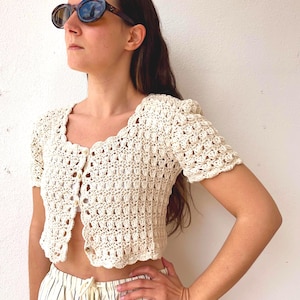 Puede incluir: Top de crochet color crema con mangas cortas y borde festoneado. El top está abotonado en la parte delantera y tiene un diseño texturizado de tejido abierto. La persona también lleva gafas de sol y pantalones a rayas.
