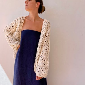 May include: A white crochet cardigan with a square stitch pattern. The cardigan is open in the front and has long sleeves. The model is wearing the cardigan over a dark blue strapless dress.