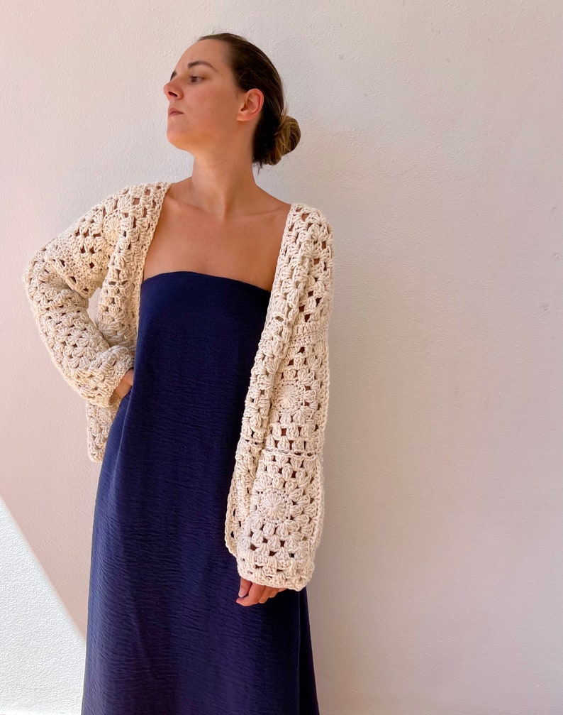 May include: A white crochet cardigan with a square stitch pattern. The cardigan is open in the front and has long sleeves. The model is wearing the cardigan over a dark blue strapless dress.