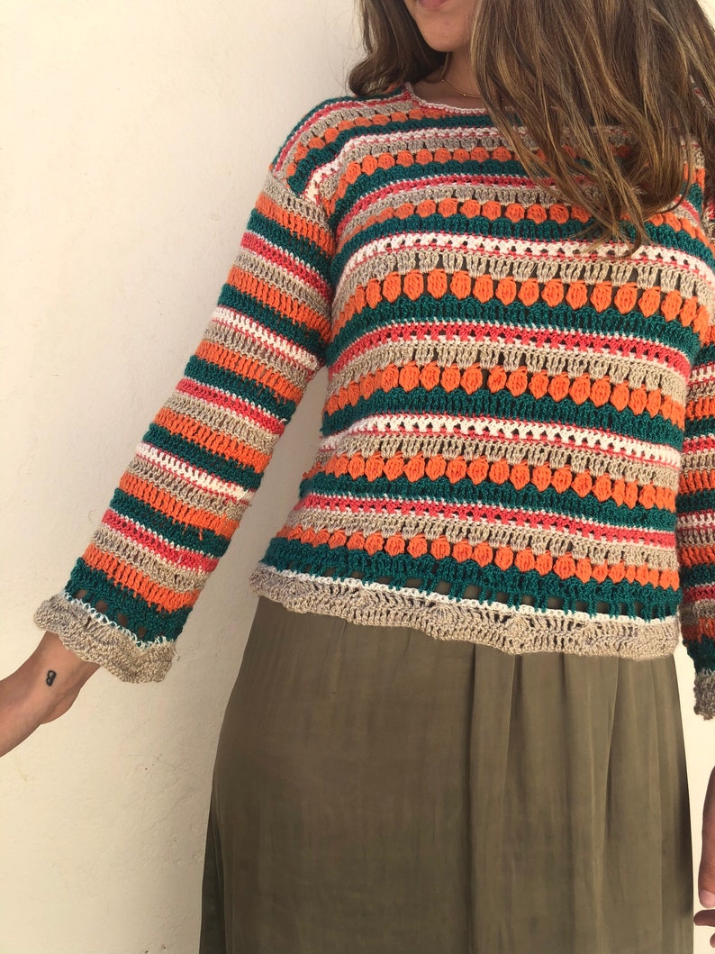 May include: A long-sleeved, striped crochet sweater with a loose fit. The sweater is made with a variety of colors, including green, orange, beige, and white.
