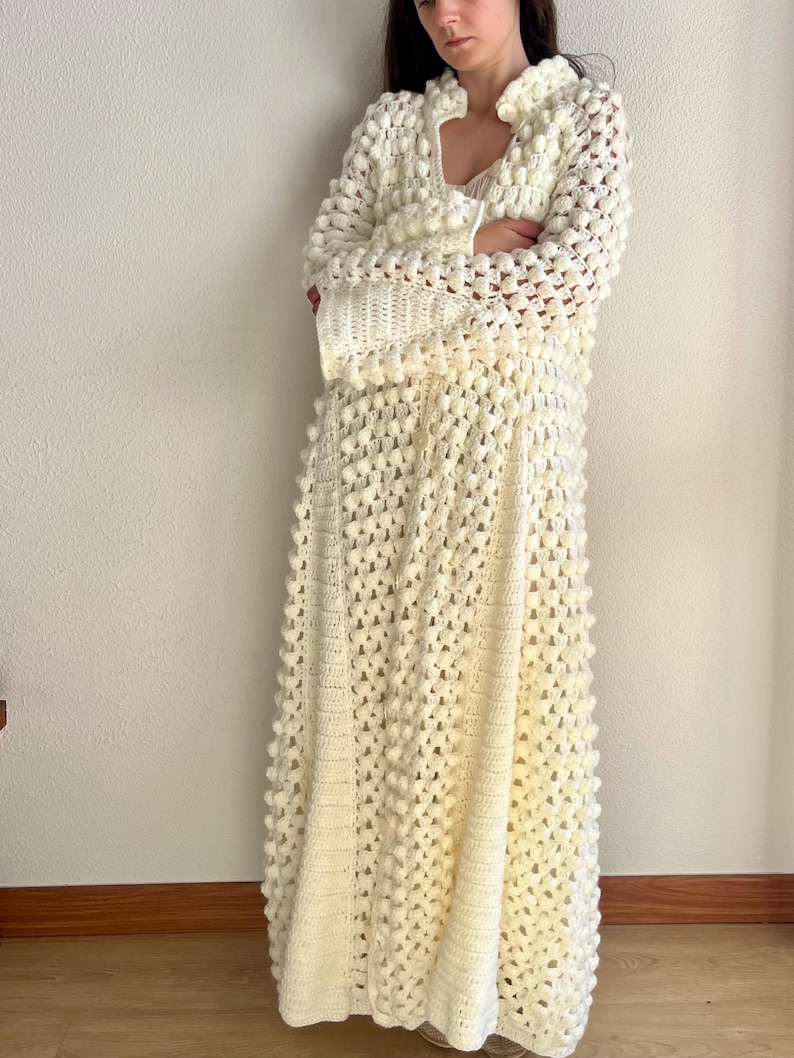 May include: A long, white crocheted dress with a textured, bobble stitch pattern. The dress has a V-neckline and long sleeves.