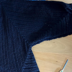 May include: A dark navy blue, handmade crochet cardigan. The cardigan features a textured stitch pattern and long sleeves. The garment is laid out on a light brown wooden surface. A pair of scissors is visible in the lower right corner.