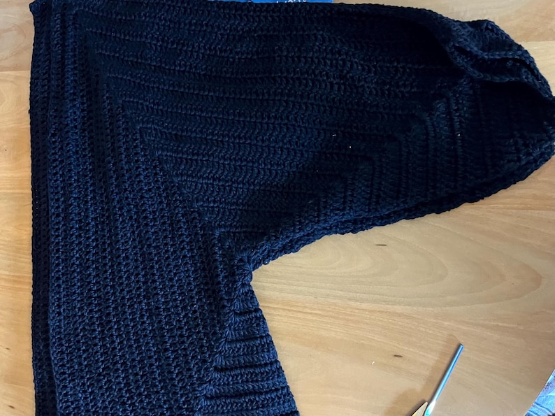May include: A dark navy blue, handmade crochet cardigan. The cardigan features a textured stitch pattern and long sleeves. The garment is laid out on a light brown wooden surface. A pair of scissors is visible in the lower right corner.
