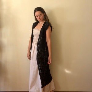 Puede incluir: Un c&aacute;rdigan de crochet negro con un frente largo abierto y sin mangas. El c&aacute;rdigan se lleva sobre un vestido blanco sin tirantes y fruncido.
