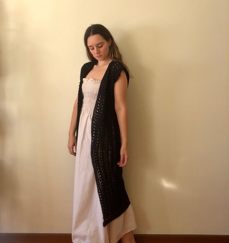Puede incluir: Un c&aacute;rdigan de crochet negro con un frente largo abierto y sin mangas. El c&aacute;rdigan se lleva sobre un vestido blanco sin tirantes y fruncido.