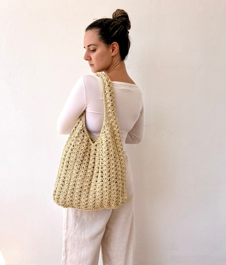 K&ouml;nnte beinhalten: Eine Frau tr&auml;gt eine hellbeige geh&auml;kelte Tragetasche &uuml;ber der Schulter. Die Tasche hat eine breite, offene Oberseite und einen langen Riemen. Die Tasche ist aus einem dicken, strukturierten Garn gefertigt.