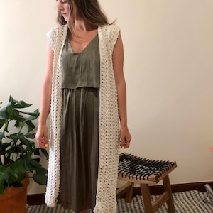 Puede incluir: Un c&aacute;rdigan de crochet blanco con un frente largo y abierto y sin mangas. El c&aacute;rdigan se lleva sobre un vestido verde sin mangas.