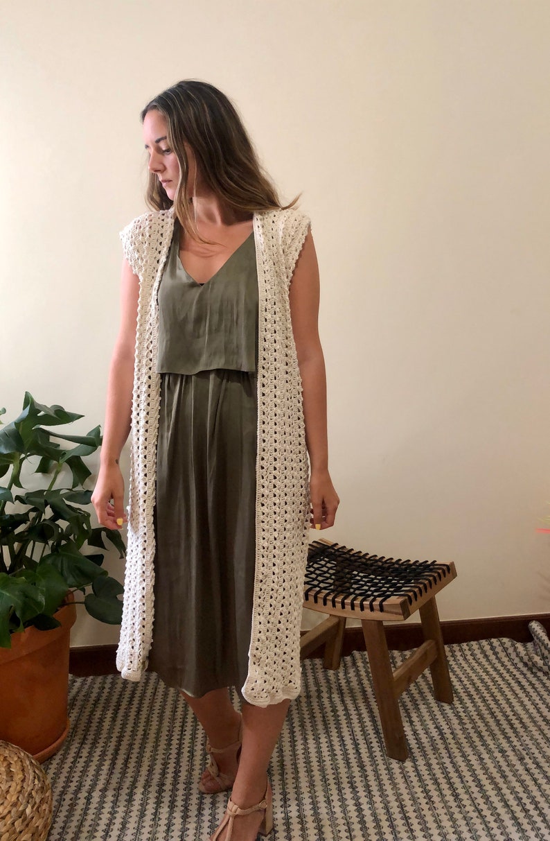Puede incluir: Un c&aacute;rdigan de crochet blanco con un frente largo y abierto y sin mangas. El c&aacute;rdigan se lleva sobre un vestido verde sin mangas.