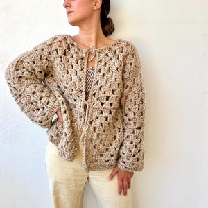 Puede incluir: Un cárdigan de crochet beige con un frente de nudo y mangas acampanadas. El cárdigan está hecho con un patrón de punto suelto y abierto.