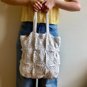 May include: A white crocheted tote bag with a square pattern. The bag is being held by a person wearing a yellow crocheted top and blue jeans.