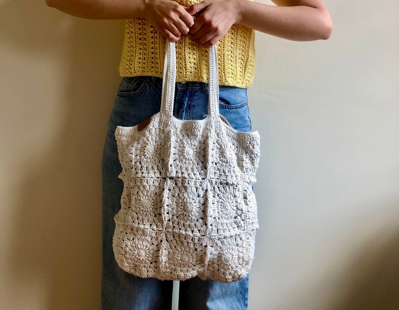 May include: A white crocheted tote bag with a square pattern. The bag is being held by a person wearing a yellow crocheted top and blue jeans.