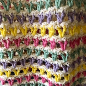 May include: Close-up of a crocheted fabric with a repeating pattern of pink, yellow, green, and purple yarn. The fabric is made with a white yarn base.