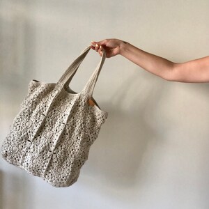 May include: A crocheted tote bag with a cream color and a square pattern. The bag has two handles and is made of a soft, textured yarn.