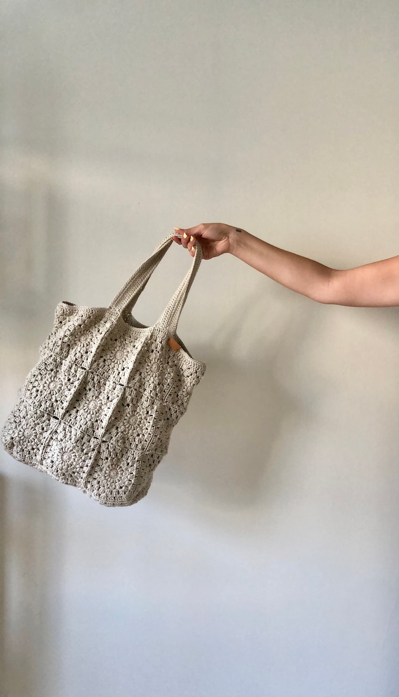 May include: A crocheted tote bag with a cream color and a square pattern. The bag has two handles and is made of a soft, textured yarn.