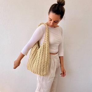 K&ouml;nnte beinhalten: Eine Person h&auml;lt eine beige geh&auml;kelte Tragetasche mit einer breiten, offenen Oberseite. Die Tasche hat eine gewebte Textur und ist aus Naturfasern gefertigt.