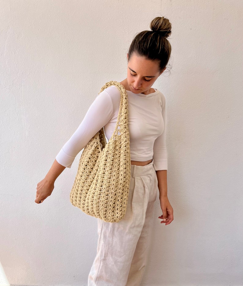 K&ouml;nnte beinhalten: Eine Person h&auml;lt eine beige geh&auml;kelte Tragetasche mit einer breiten, offenen Oberseite. Die Tasche hat eine gewebte Textur und ist aus Naturfasern gefertigt.