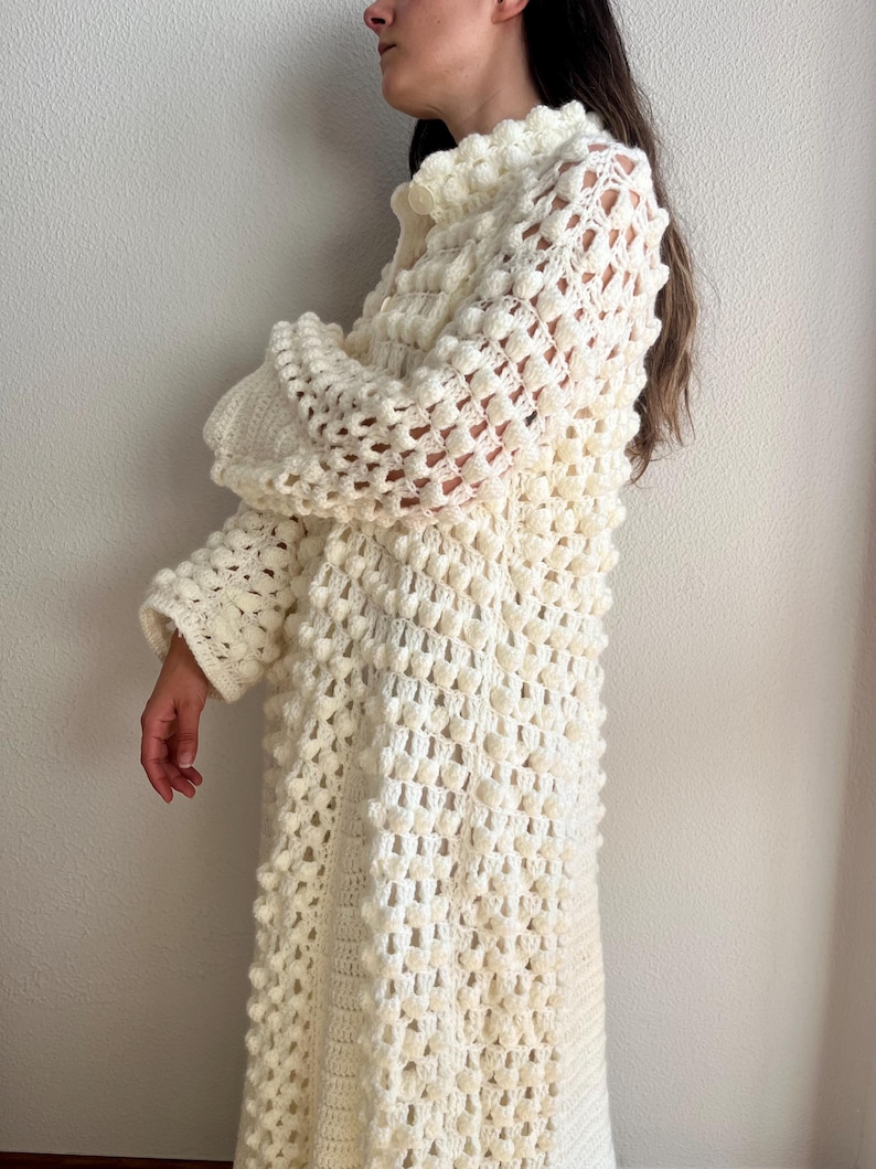May include: A white crocheted long-sleeved dress with a textured, bobble stitch pattern. The dress has a high neckline and a button closure.