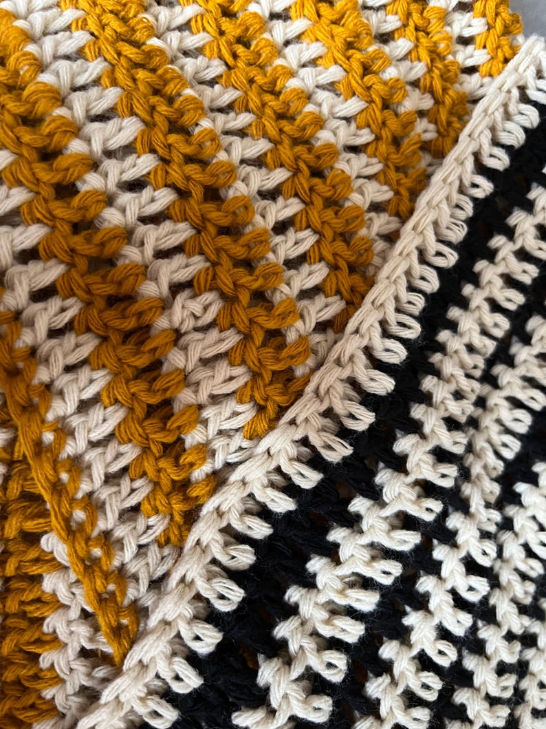 May include: Close-up of a crocheted textile with a geometric pattern. The design features sections of mustard yellow, cream, and black, creating a textured and visually appealing surface. The crochet stitches are clearly defined.