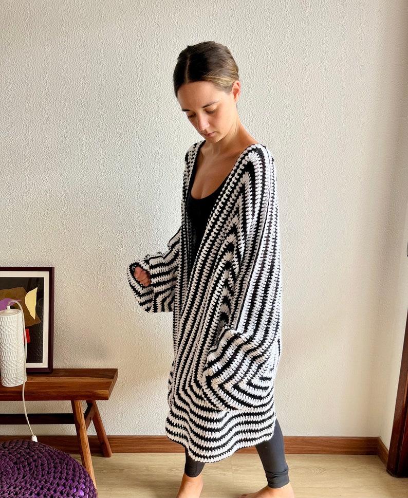 May include: A long, open-front crochet cardigan in a bold black and white striped pattern. The handmade garment features a relaxed fit with wide sleeves and a knee-length hem. The cardigan is worn over a black top and leggings.