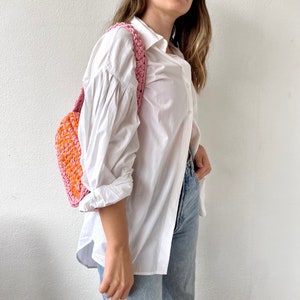 May include: A woman wearing a white button-down shirt and blue jeans is carrying a pink and orange crocheted bag.
