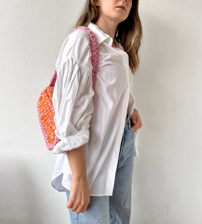 May include: A woman wearing a white button-down shirt and blue jeans is carrying a pink and orange crocheted bag.