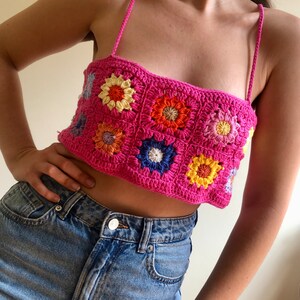 May include: A pink crochet top with a floral pattern. The top is made of granny squares with colorful flowers in the center. The top is sleeveless and has a square neckline.