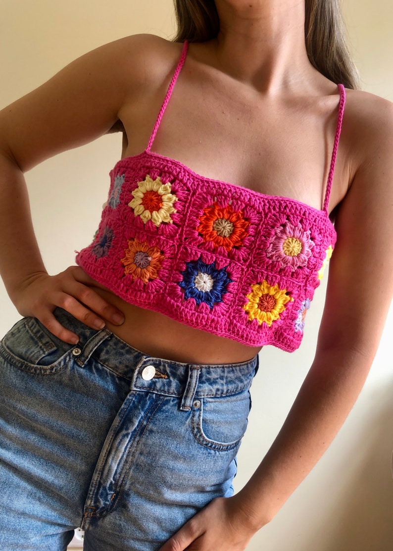 May include: A pink crochet top with a floral pattern. The top is made of granny squares with colorful flowers in the center. The top is sleeveless and has a square neckline.