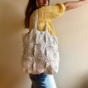 May include: A white crocheted tote bag with a square pattern. The bag has two long straps and is being held by a person wearing a yellow crocheted top and blue jeans.