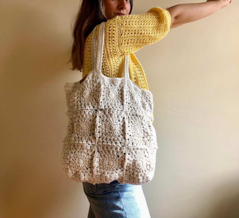 May include: A white crocheted tote bag with a square pattern. The bag has two long straps and is being held by a person wearing a yellow crocheted top and blue jeans.