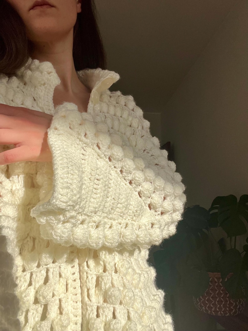 May include: A white crocheted cardigan with a textured design and pom-pom accents.