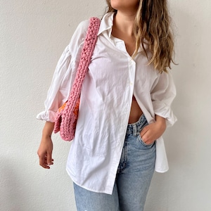 May include: A woman wearing a white oversized button-down shirt with a pink and orange crocheted bag slung over her shoulder. She is wearing light blue jeans and has her hand in her pocket.