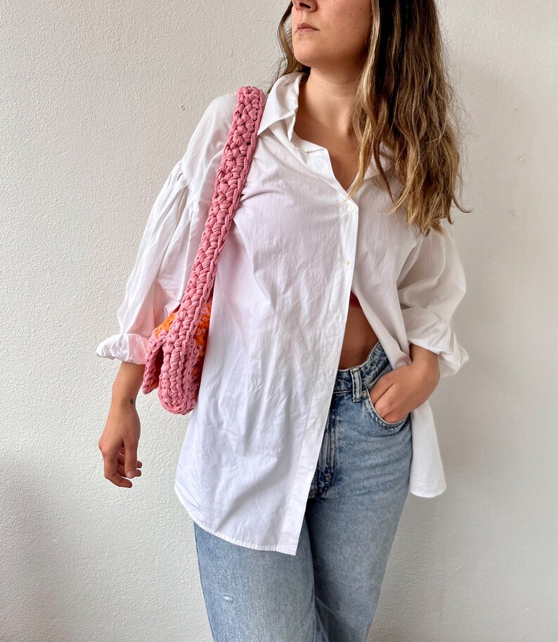 May include: A woman wearing a white oversized button-down shirt with a pink and orange crocheted bag slung over her shoulder. She is wearing light blue jeans and has her hand in her pocket.