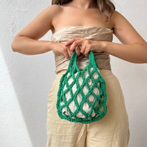 May include: A green crocheted mesh bag with a white fabric lining. The bag is being held by a person.