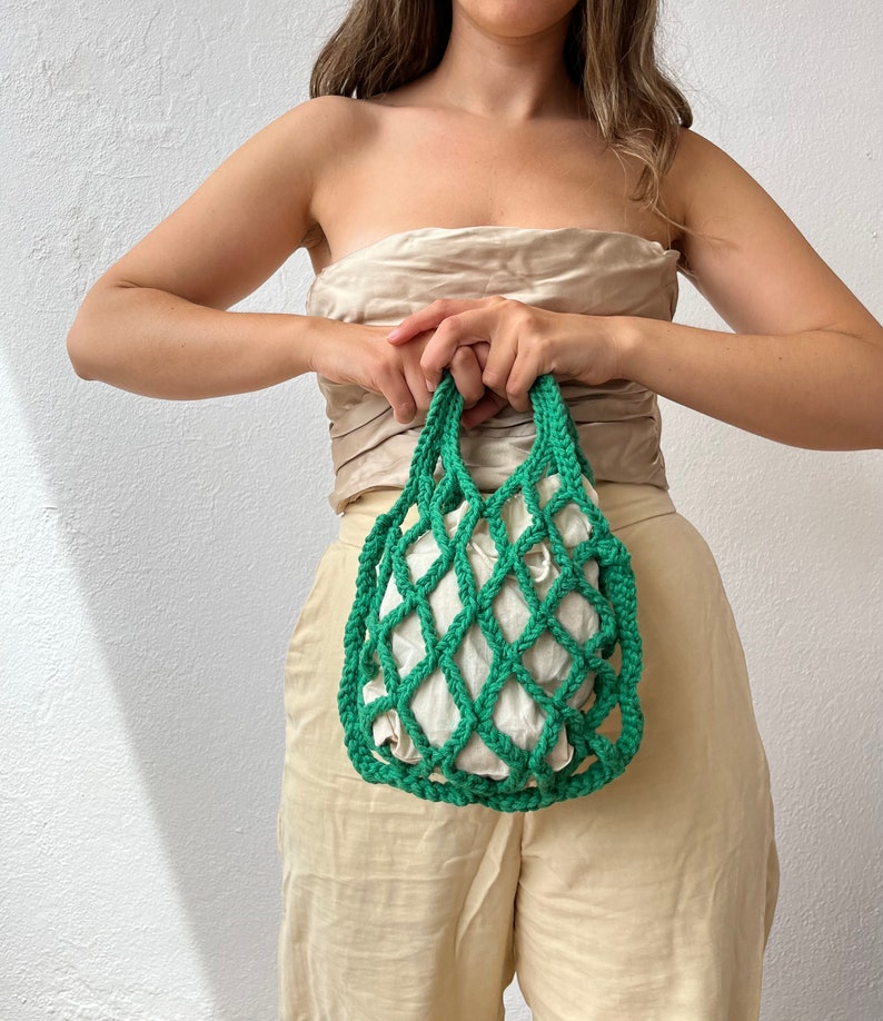 May include: A green crocheted mesh bag with a white fabric lining. The bag is being held by a person.