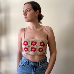 May include: A crocheted crop top with a geometric pattern of red and beige squares. The top has thin straps and a relaxed fit.