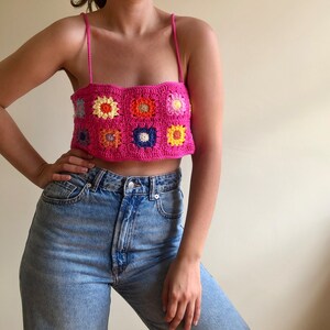 K&ouml;nnte beinhalten: Ein leuchtend pinkes H&auml;keltop mit floralem Design. Das Top hat ein quadratisches Muster mit bunten Blumenmotiven in Gelb, Orange, Blau und Rosa. Es wird mit hellblauen Jeans kombiniert.
