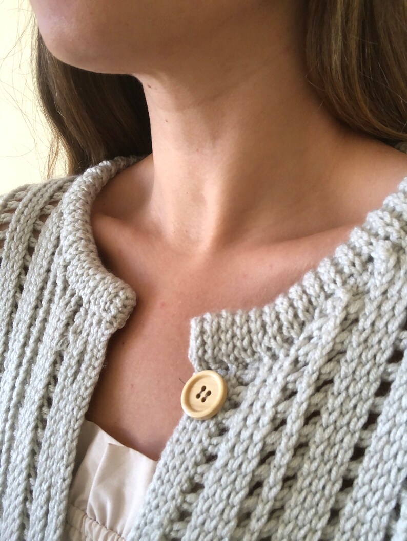 K&ouml;nnte beinhalten: Nahaufnahme eines hellgrauen Strickjackens mit einem Holzknopf. Der Cardigan hat eine lockere, offene Maschenstruktur.