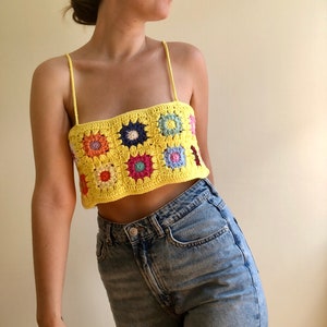 May include: A yellow crochet top with a colorful floral pattern. The top is sleeveless and has a square neckline. The top is cropped and shows the wearer's midriff.