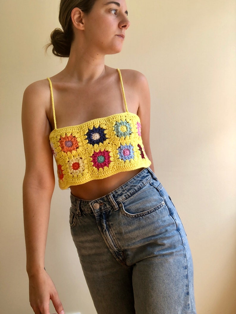 May include: A yellow crochet top with a colorful floral pattern. The top is sleeveless and has a square neckline. The top is cropped and shows the wearer's midriff.