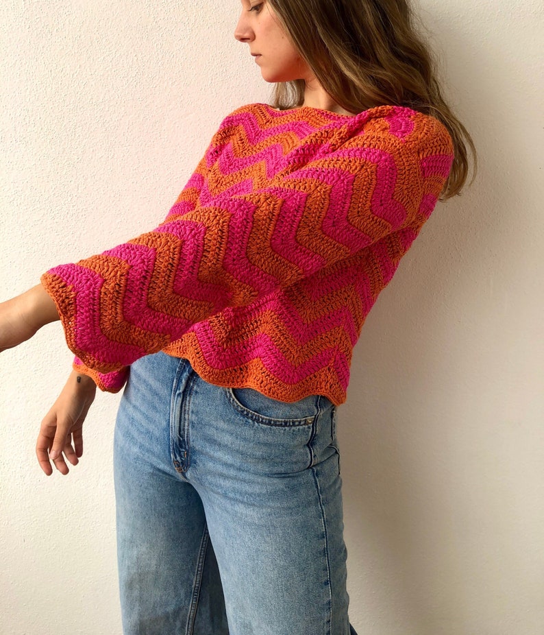 May include: A pink and orange crochet sweater with a chevron pattern. The sweater has long sleeves and a relaxed fit.
