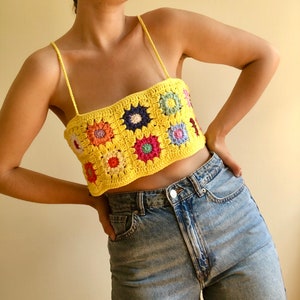 May include: A yellow crochet top with colorful flower accents. The top is sleeveless and has a square neckline. The model is wearing blue jeans.