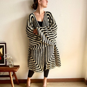 May include: A long, open-front crochet cardigan in a striking black and cream geometric pattern. The cardigan has wide sleeves and a knee-length hem. The person is wearing black leggings and gold-colored square-toe shoes.