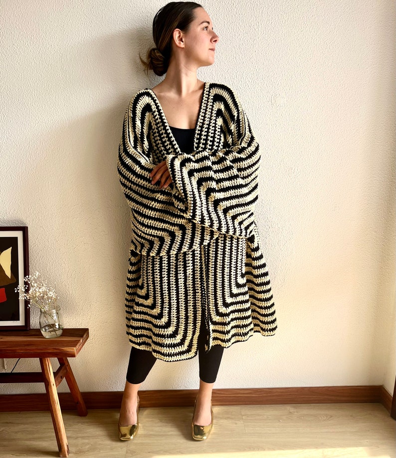 May include: A long, open-front crochet cardigan in a striking black and cream geometric pattern. The cardigan has wide sleeves and a knee-length hem. The person is wearing black leggings and gold-colored square-toe shoes.