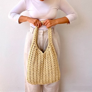 K&ouml;nnte beinhalten: Eine beige geh&auml;kelte Tragetasche mit einer breiten, offenen Oberseite und einem einzigen Henkel. Die Tasche ist aus dickem, strukturiertem Garn gefertigt und hat eine leicht schlaffe Form.