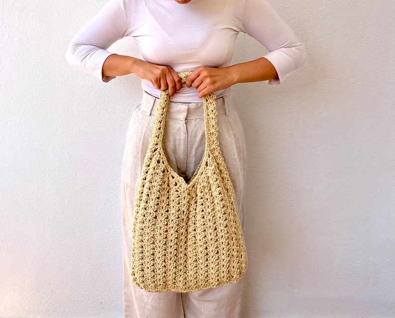 K&ouml;nnte beinhalten: Eine beige geh&auml;kelte Tragetasche mit einer breiten, offenen Oberseite und einem einzigen Henkel. Die Tasche ist aus dickem, strukturiertem Garn gefertigt und hat eine leicht schlaffe Form.