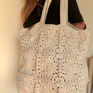 May include: A large, off-white crocheted tote bag with a square pattern. The bag has two thick straps and is made of a soft, textured yarn.