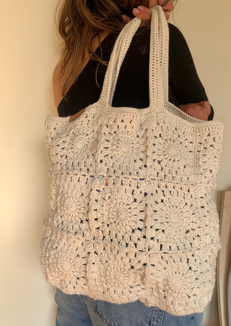 May include: A large, off-white crocheted tote bag with a square pattern. The bag has two thick straps and is made of a soft, textured yarn.