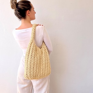 K&ouml;nnte beinhalten: Eine Frau in einem wei&szlig;en Shirt und wei&szlig;en Hosen tr&auml;gt eine gro&szlig;e, geh&auml;kelte, beige Tasche &uuml;ber der Schulter.