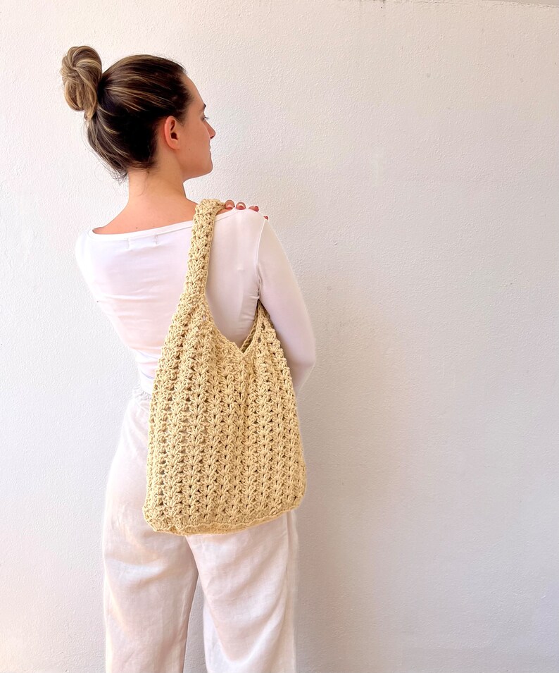 K&ouml;nnte beinhalten: Eine Frau in einem wei&szlig;en Shirt und wei&szlig;en Hosen tr&auml;gt eine gro&szlig;e, geh&auml;kelte, beige Tasche &uuml;ber der Schulter.
