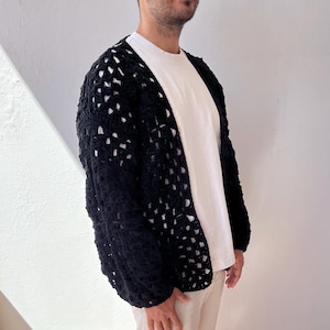 K&ouml;nnte beinhalten: Ein schwarzer H&auml;kelcardigan mit quadratischem Muster. Der Cardigan ist vorne offen und hat lange &Auml;rmel.